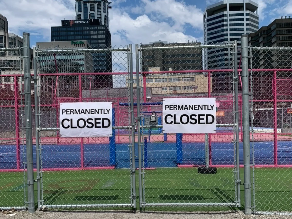 Chain link gates with "permanently closed" sign in front of blue and green court surfaces with tall buildings in background.