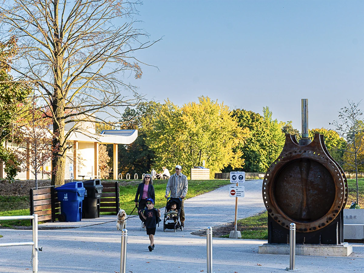 Family with stroller on wide paved walkway with park pavilion and trees in background