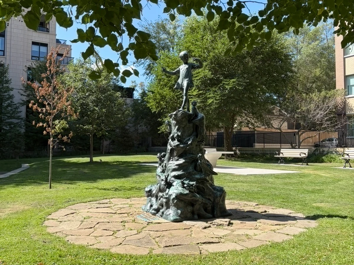 Bronze statue of small boy on small round stone-paved area surrounded by grass and trees.